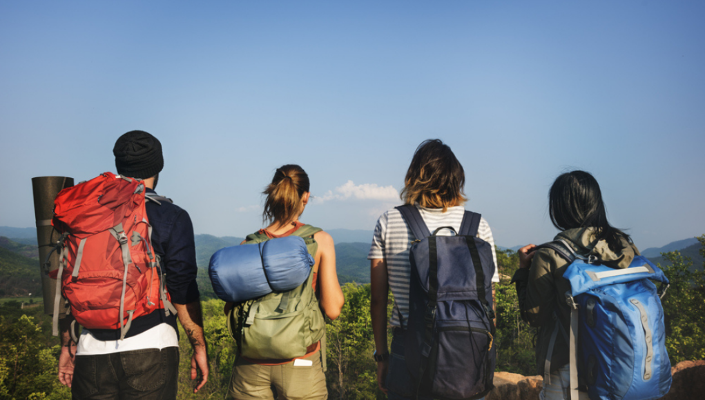 young-adventure-travellers-backpacking-green-hospitality Four young adventure travellers with large backpacks and hiking gear standing on a ridge overlooking a vast green valley under a clear blue sky.