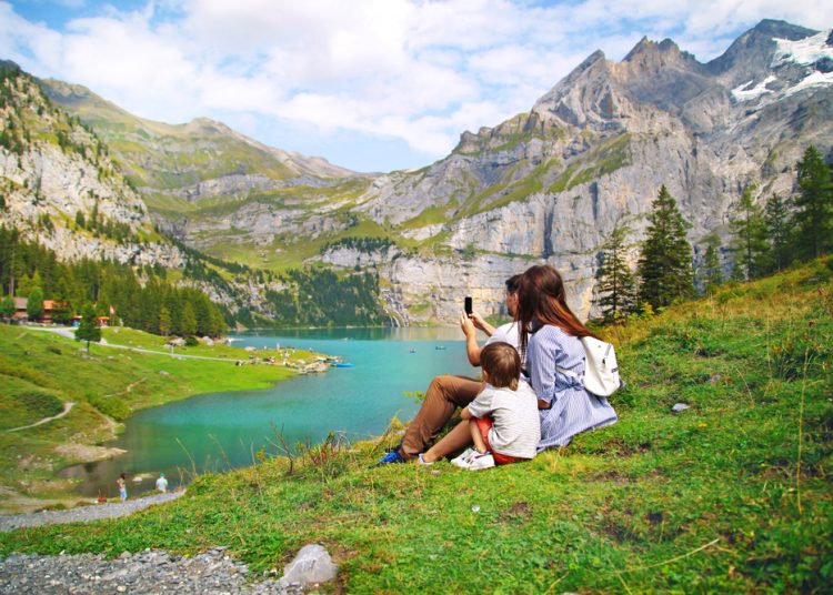 Adventure Tourism-How Hospitality Can Win Travellers with Scuffed Boots and Green Hearts 8 A family sitting on a grassy hillside overlooking a crystal-clear turquoise alpine lake surrounded by dramatic, snow-capped mountains in a premier eco-tourism destination.