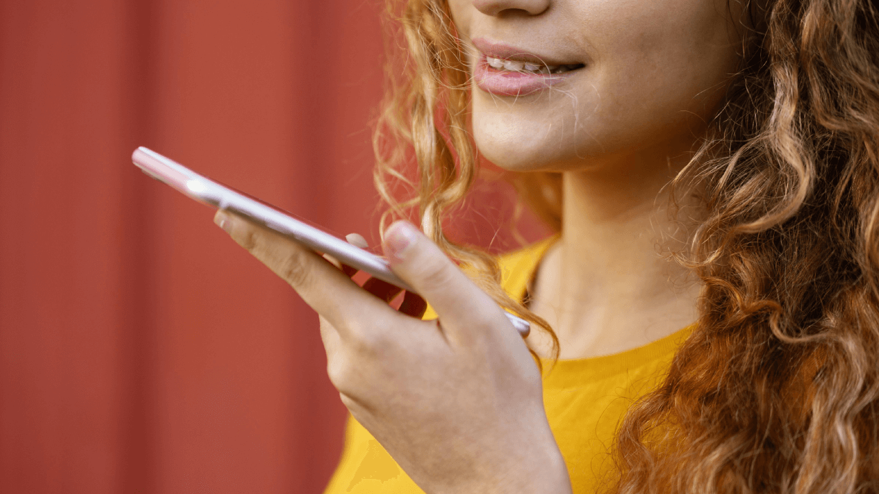 "A young woman with curly hair speaking into her smartphone, using voice search against a vibrant red background."
